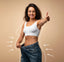 Woman in a white sports bra and blue jeans showing off weight loss against a beige background